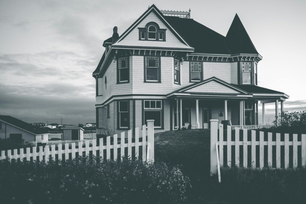 A classic Victorian house with a white picket fence in a rural area, captured in black and white.